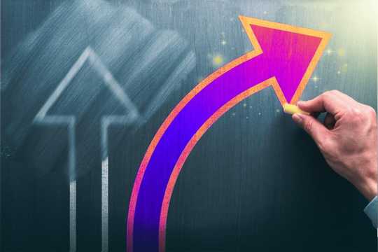 Value shifts. A hand draws a bright, curved neon-pink and orange arrow on a dark chalkboard, rising upward beside a faint, blurred white arrow in the background.