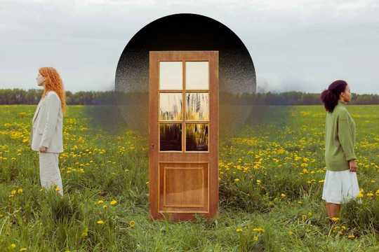 Structural dissociation. Two people stand facing away from each other in a green field of yellow flowers, separated by a central wooden door framed by a dark arch, creating a symmetrical, surreal composition in soft natural greens, yellows, browns, and muted neutrals.
